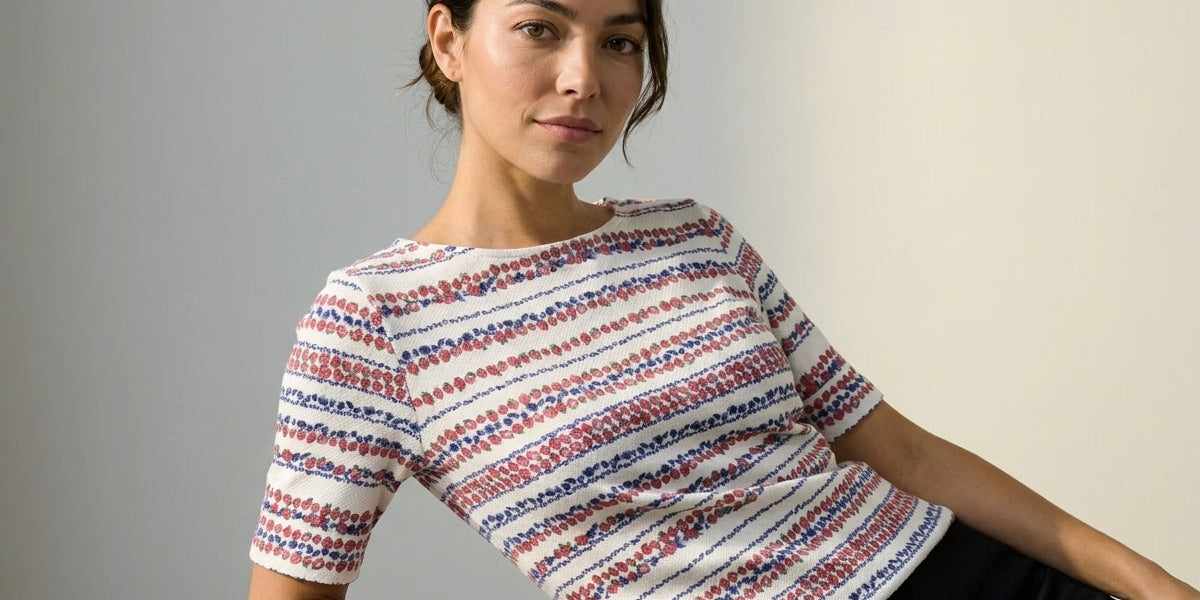 A woman wearing a short-sleeve knit top with horizontal red, blue, and white textured stripes, leaning on a light gray block against a neutral studio background.