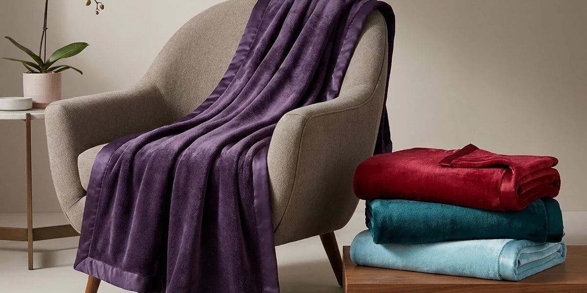 Plush purple fleece blanket with satin trim draped over a modern armchair, next to a stack of folded red, teal, and light blue blankets.