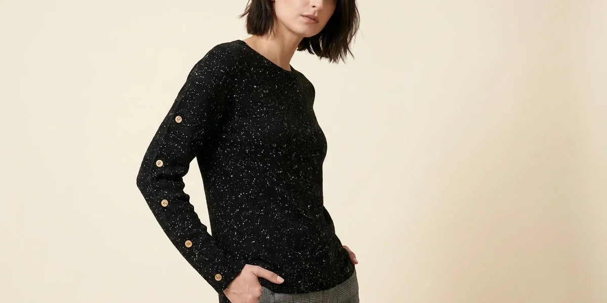 Brunette woman modeling a black speckled knit crewneck sweater with button accents down the sleeves, paired with grey plaid trousers.