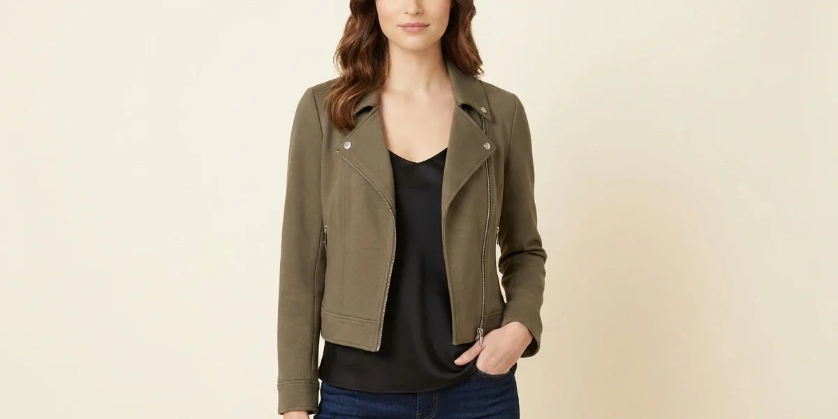A woman modeling a cropped olive green moto jacket over a black top and blue jeans, standing against a plain beige studio background.