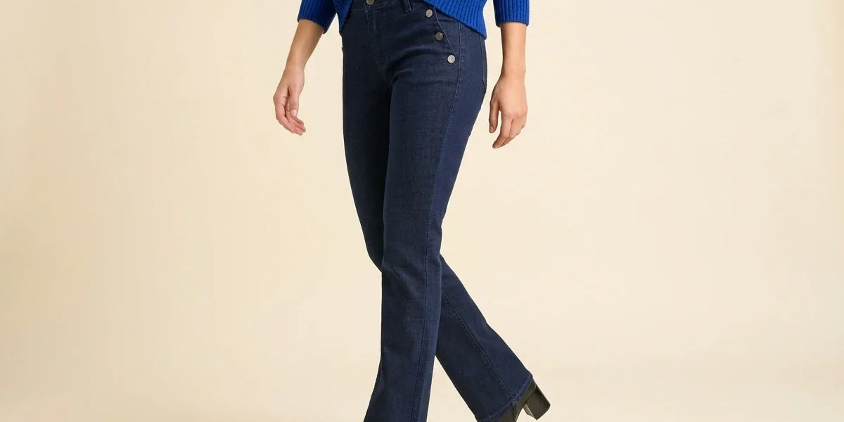 A model wearing dark blue flared jeans, a blue knit sweater, and black ankle boots, shown from the chest down against a solid beige studio background.