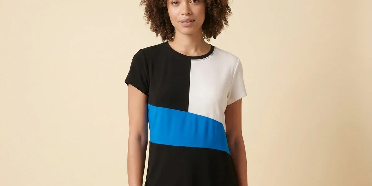 A woman with curly brown hair wearing a short-sleeve colorblock top with black, white, and bright blue blocks, paired with black pants, standing against a solid beige background.