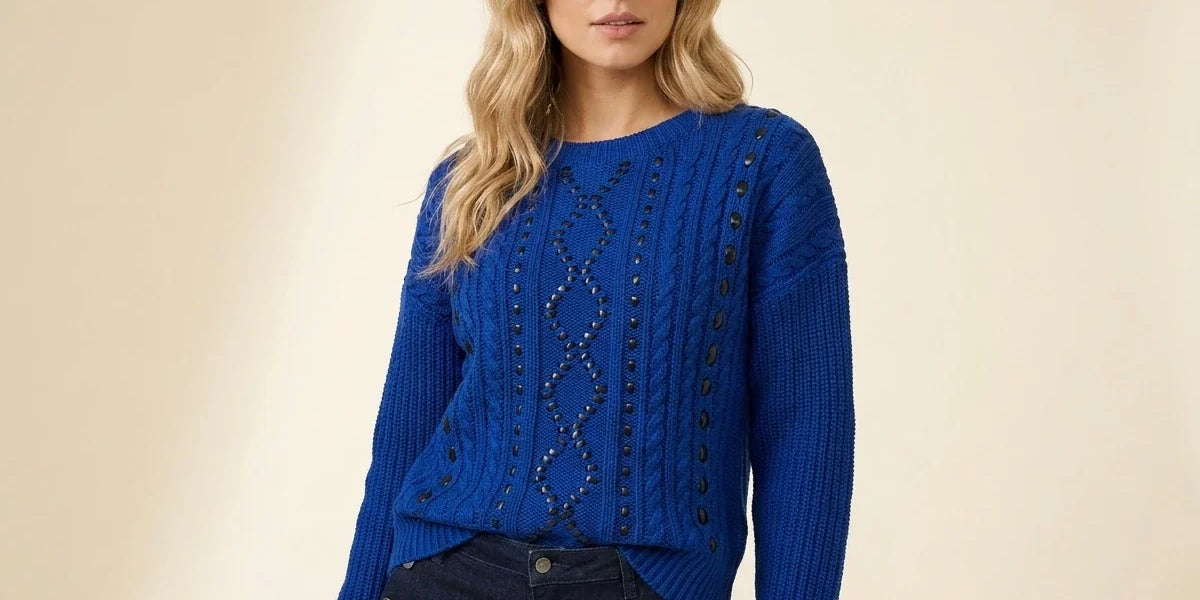 A woman modeling a royal blue cable-knit sweater with decorative dark beadwork down the center front and dark buttons along the sleeves, paired with dark jeans against a warm beige studio background.