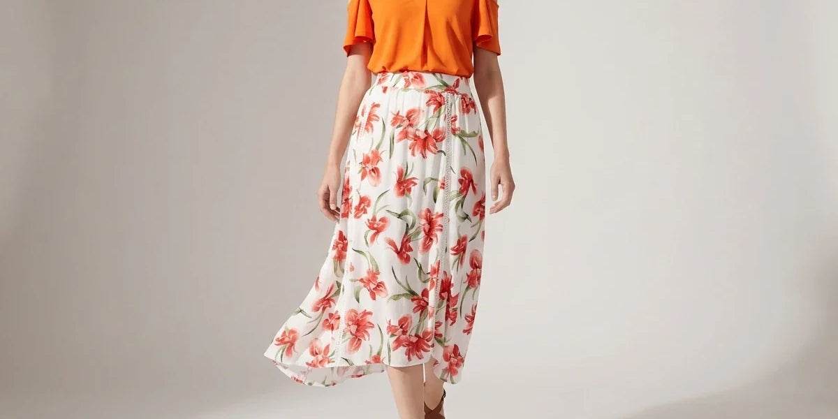 A woman modeling a two-piece outfit consisting of a bright orange cold-shoulder V-neck top and a white midi skirt with a red and green floral print, set against a plain light gray studio background.