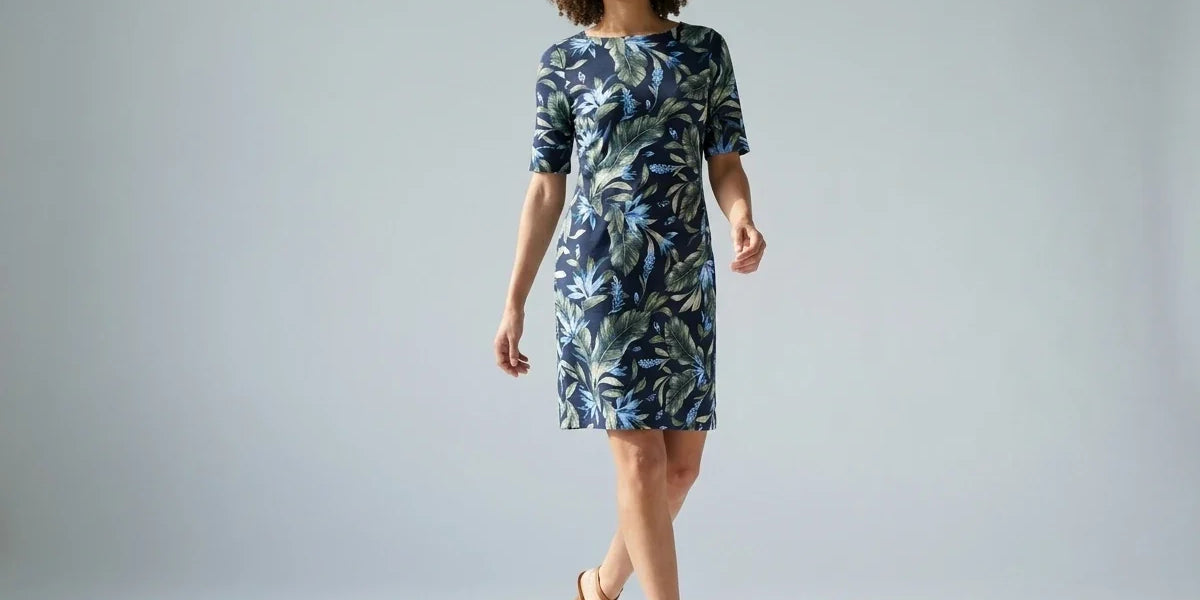 A woman modeling a knee-length short-sleeve dress with a blue and green tropical leaf pattern, paired with tan strappy high-heeled sandals, against a light gray studio background.