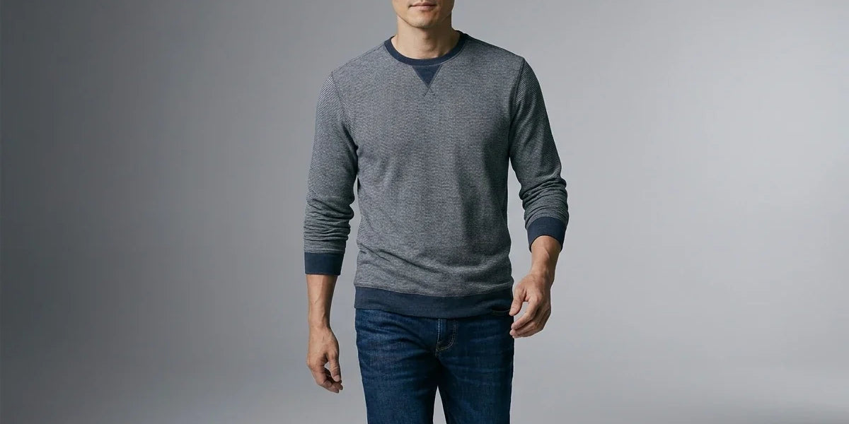 A man wearing a gray long-sleeve crewneck sweater with dark navy trim and blue jeans, standing against a neutral gray studio background.