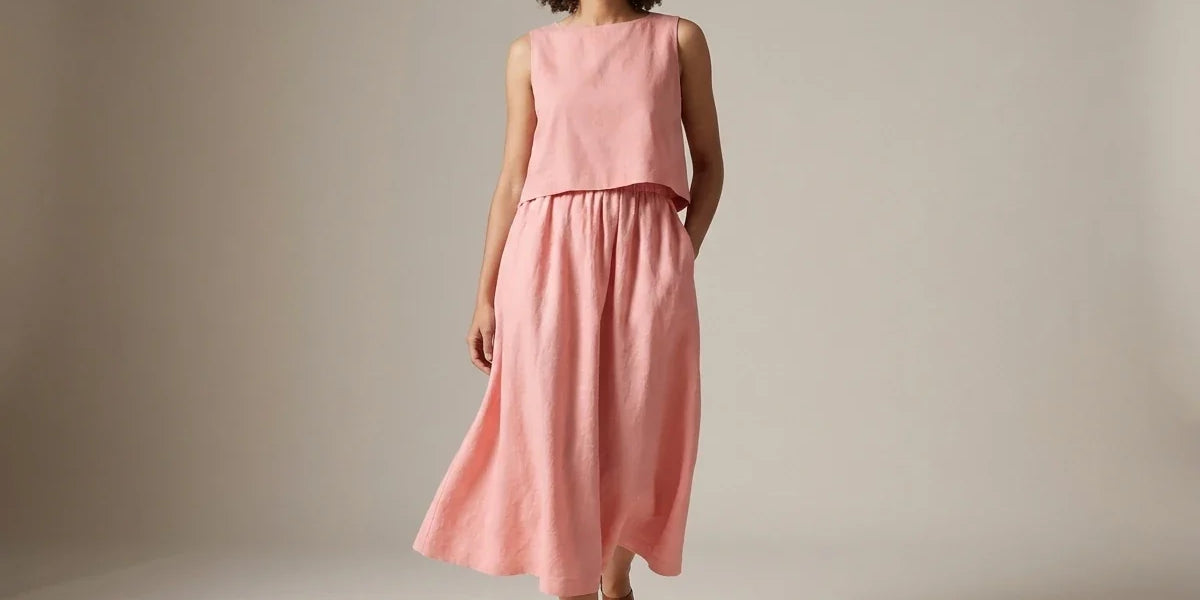 Woman with curly hair modeling a blush pink sleeveless two-piece linen midi dress and brown flat sandals in a studio setting.