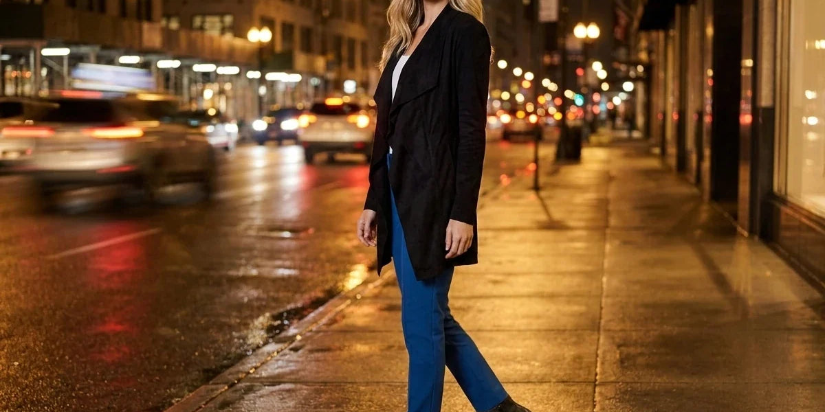 Blonde woman wearing a black drape-front trench coat, bright blue trousers, and black heeled ankle boots walking along a city sidewalk at night.