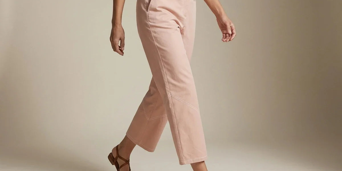 Cropped view of a woman wearing dusty pink straight-leg ankle pants and strappy brown flat sandals against a neutral studio backdrop.