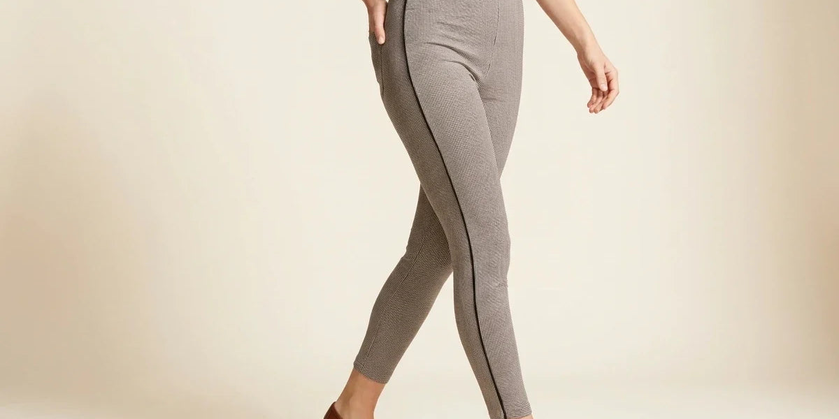 A model wearing light gray heathered leggings with a thin black side stripe, paired with a brown sleeveless top and brown leather loafers, shown from the waist down against a warm beige studio background.
