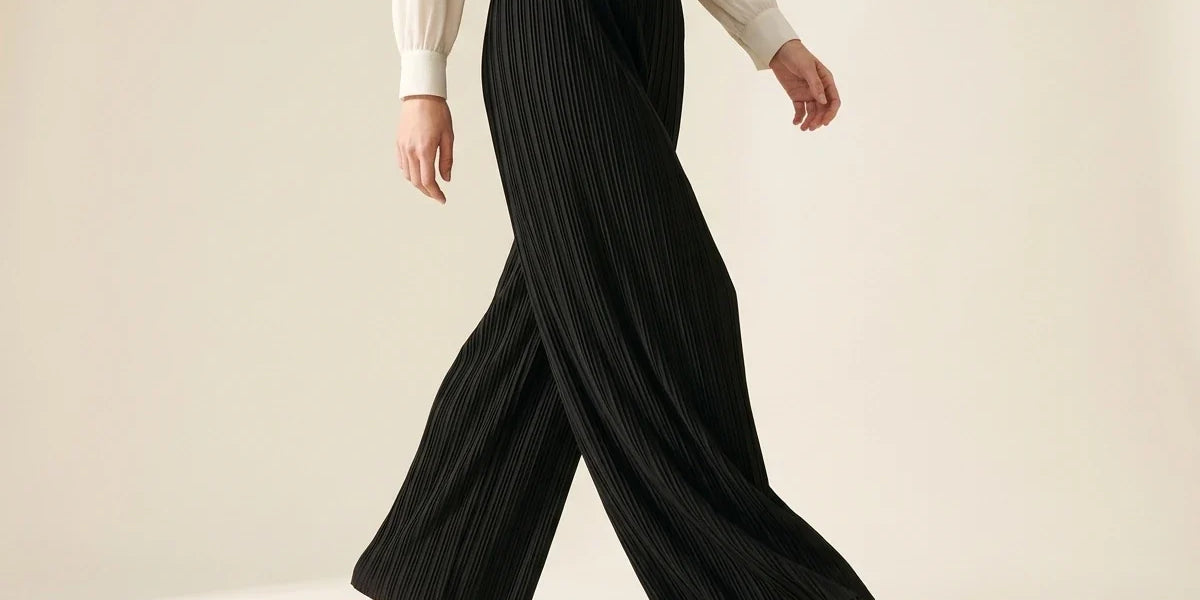 Cropped view of a woman walking in black pleated wide-leg palazzo pants paired with black pointed-toe kitten heel mules and a cream blouse.