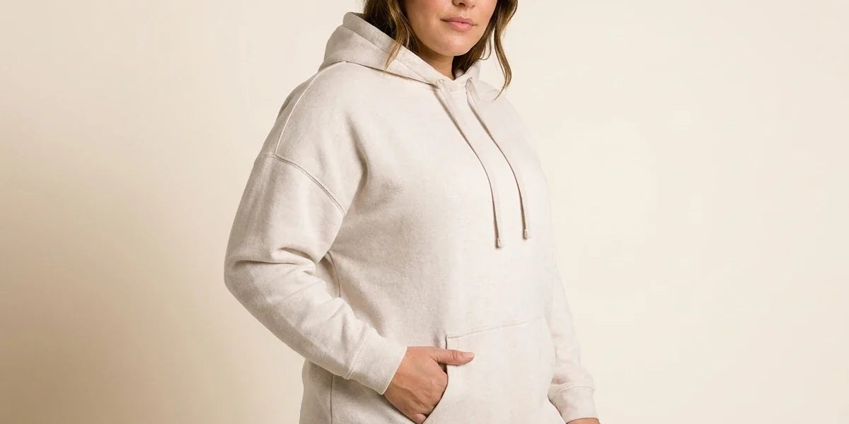 Brunette woman modeling a plus-size oatmeal heather fleece hoodie sweater with a front kangaroo pocket and black leggings.