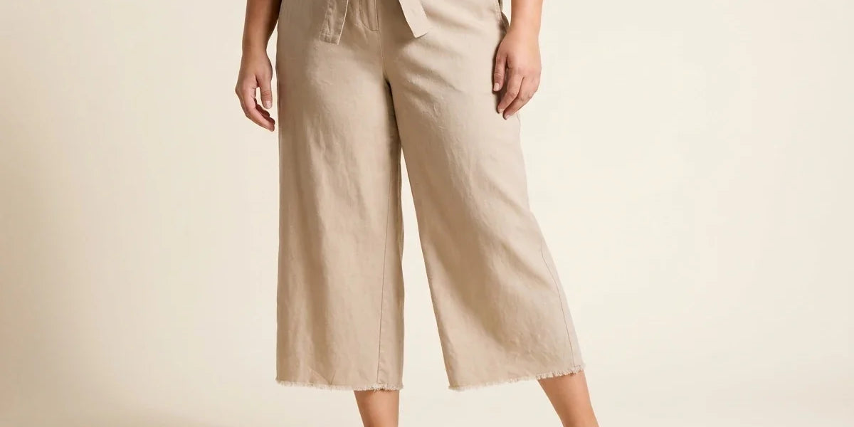Cropped view of a woman wearing high-waisted beige linen tie-front wide-leg culottes with raw fringe hems and tan leather mules.