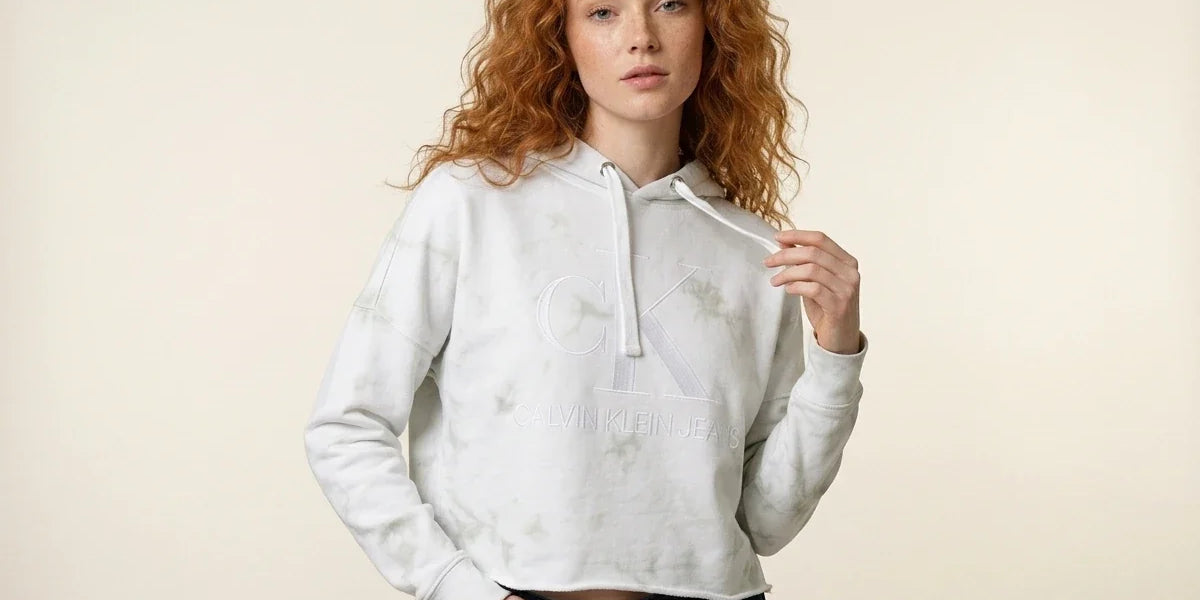 Woman with curly red hair modeling a Calvin Klein Jeans light green tie-dye cropped hoodie paired with dark wash jeans.