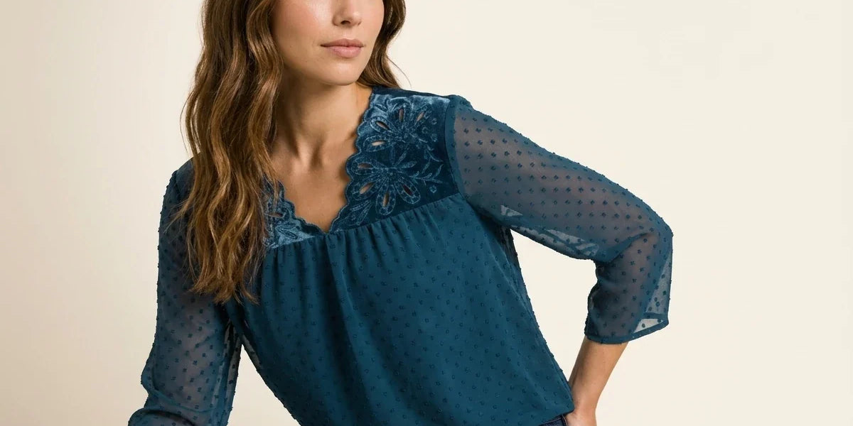Brunette woman modeling a teal blue Swiss dot chiffon blouse featuring a velvet floral cutout neckline paired with dark wash jeans.