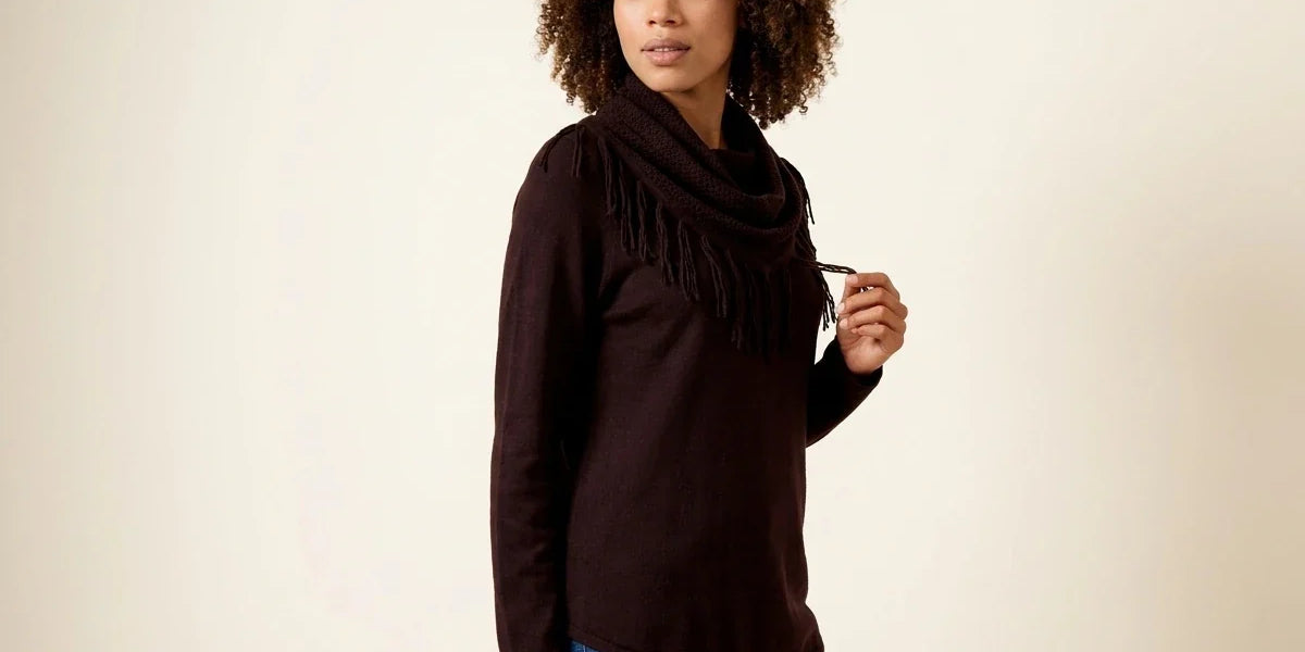 Woman with curly brown hair modeling a dark chocolate brown cowl-neck sweater with fringe trim paired with blue jeans.