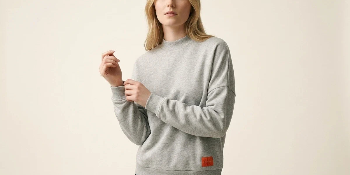 Blonde woman modeling a Calvin Klein heather grey drop-shoulder sweatshirt with an orange logo patch, paired with dark wash jeans.