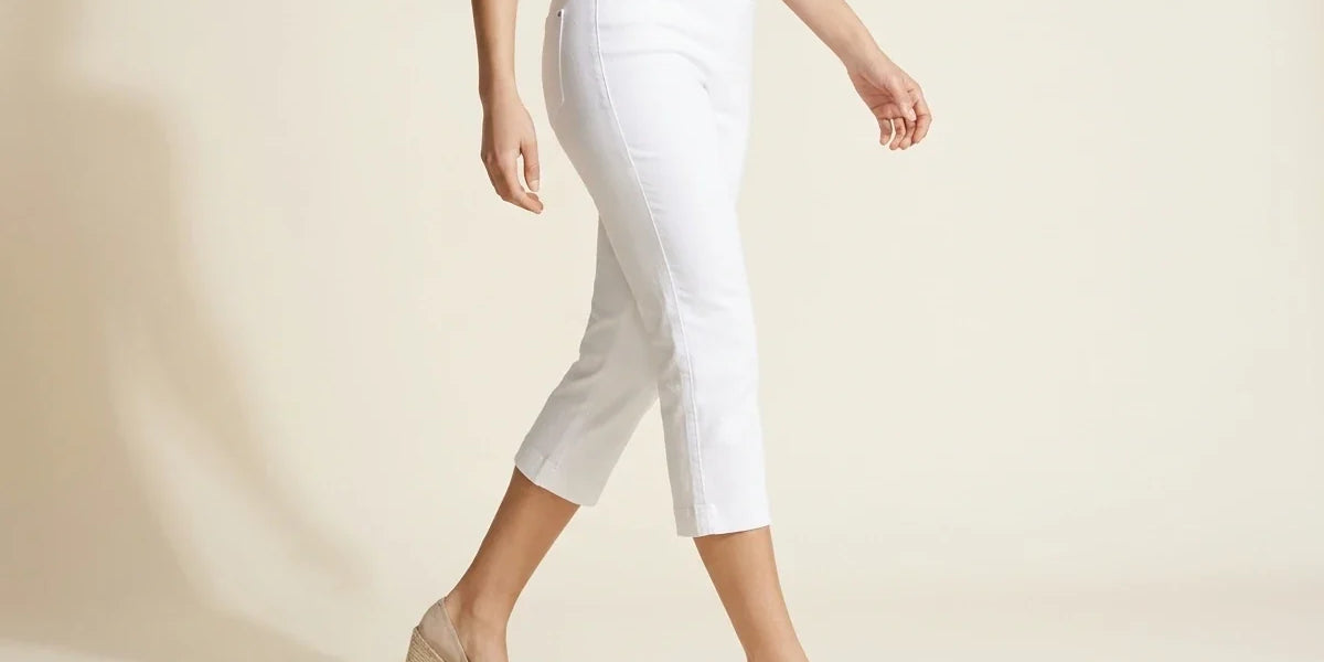 A model wearing white cropped capri pants, a light pink top, and beige wedge espadrilles, walking against a warm beige studio background.
