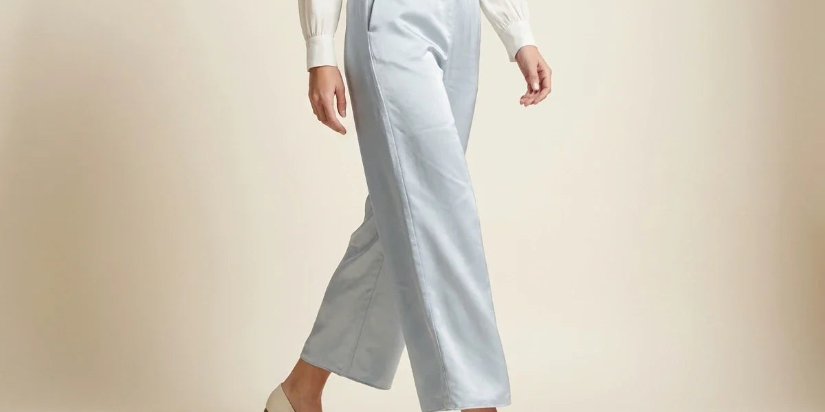 A model wearing light blue wide-leg cropped trousers, a white long-sleeve top, and beige flats, shown from the waist down against a warm beige studio background.