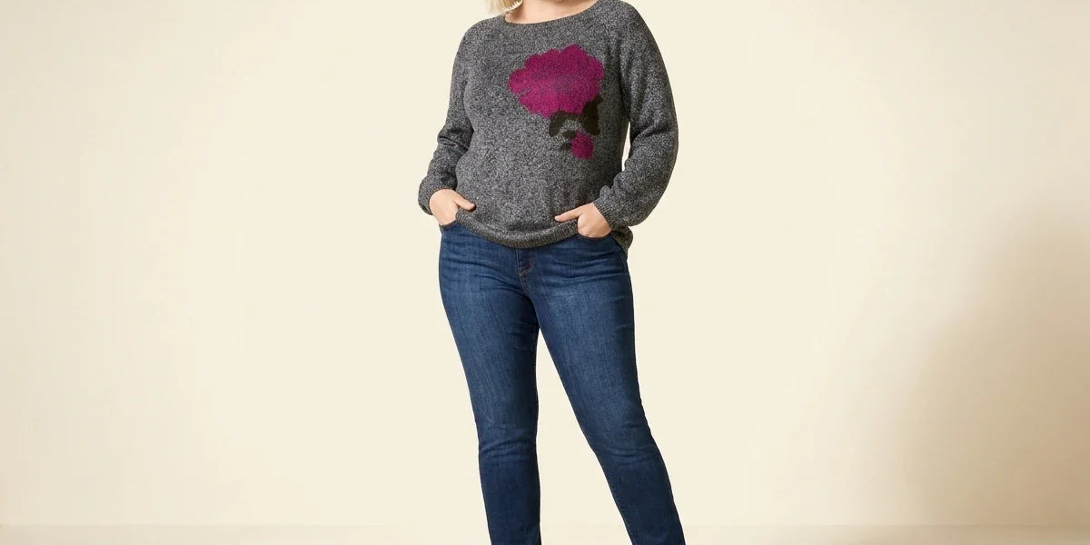 Blonde woman modeling a plus-size charcoal heather grey knit sweater with a magenta floral graphic, medium-wash skinny jeans, and brown leather ankle boots.