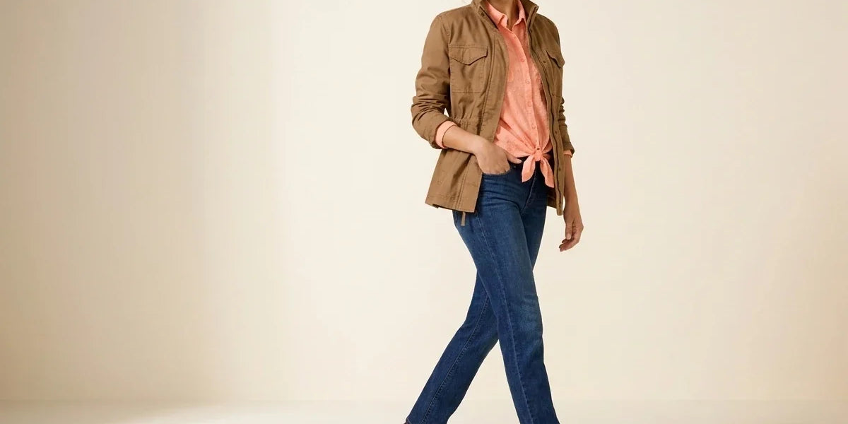 Brunette woman modeling petite-fit straight leg blue jeans with a tan utility jacket, peach tie-front shirt, and dark brown leather boots.