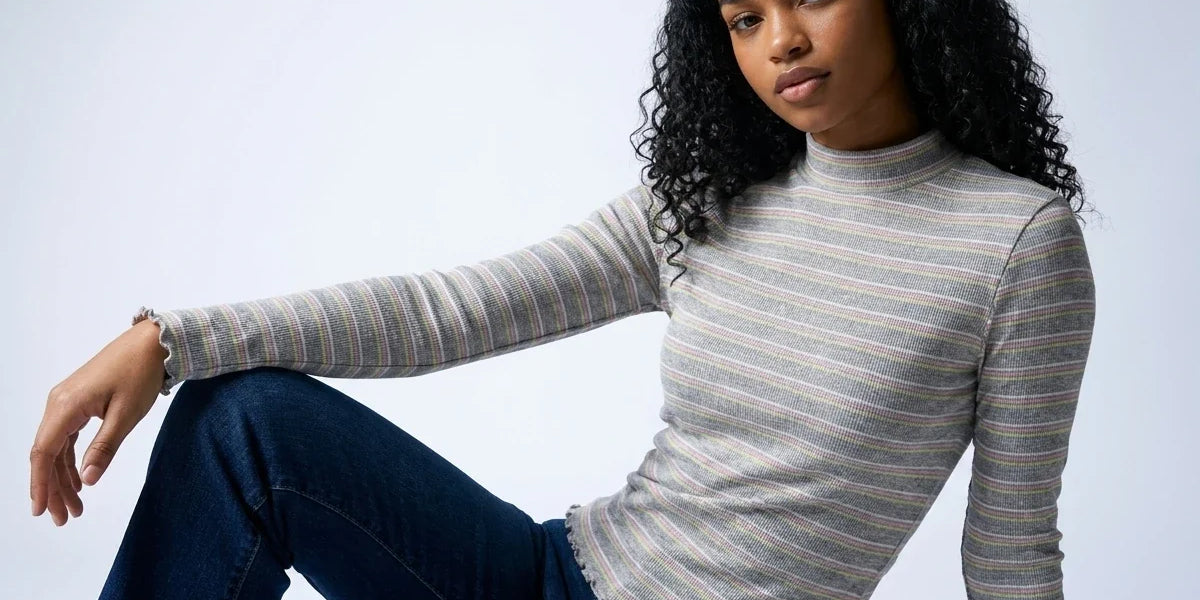 A woman with curly black hair wearing a gray and white striped mock neck long-sleeve top and dark blue jeans, posing against a light gray studio background.
