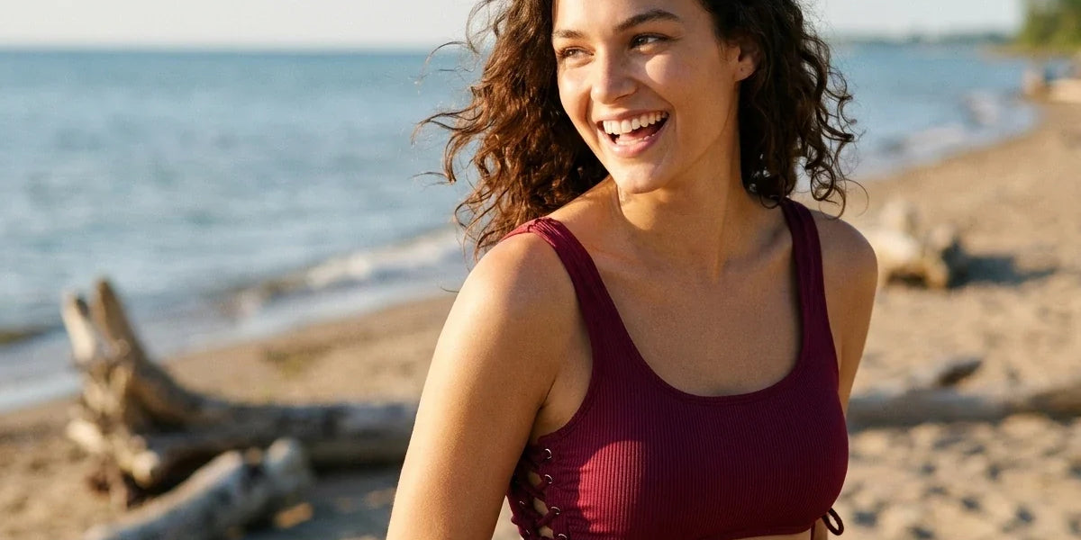 A woman with curly brown hair smiling on a sandy beach wearing a ribbed burgundy side-tie bikini top. The background shows the blue water and sky of a lake with driftwood logs on the shore.