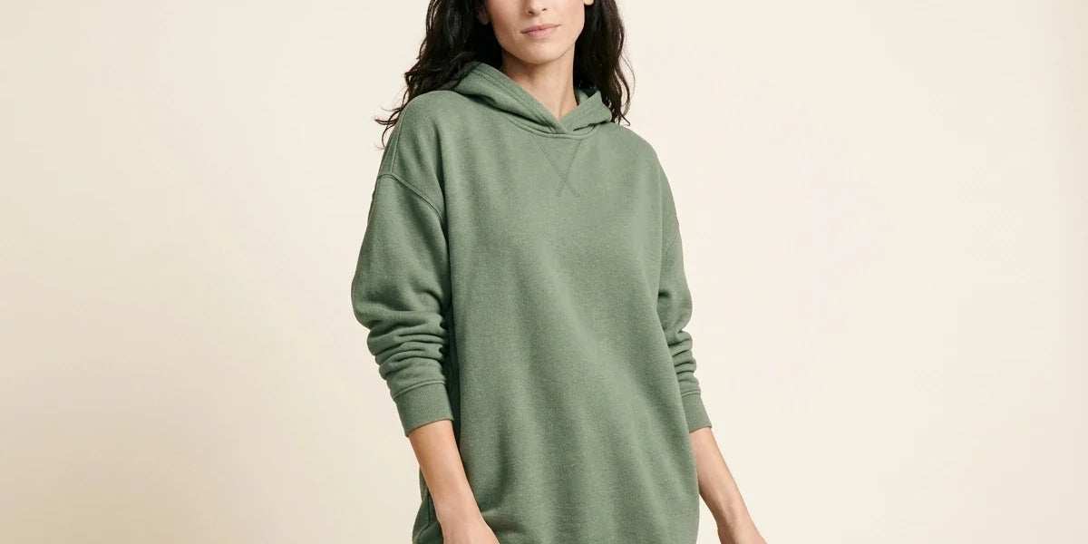 Brunette woman modeling an oversized sage green fleece hoodie sweater with cross-stitch collar detailing and black leggings.
