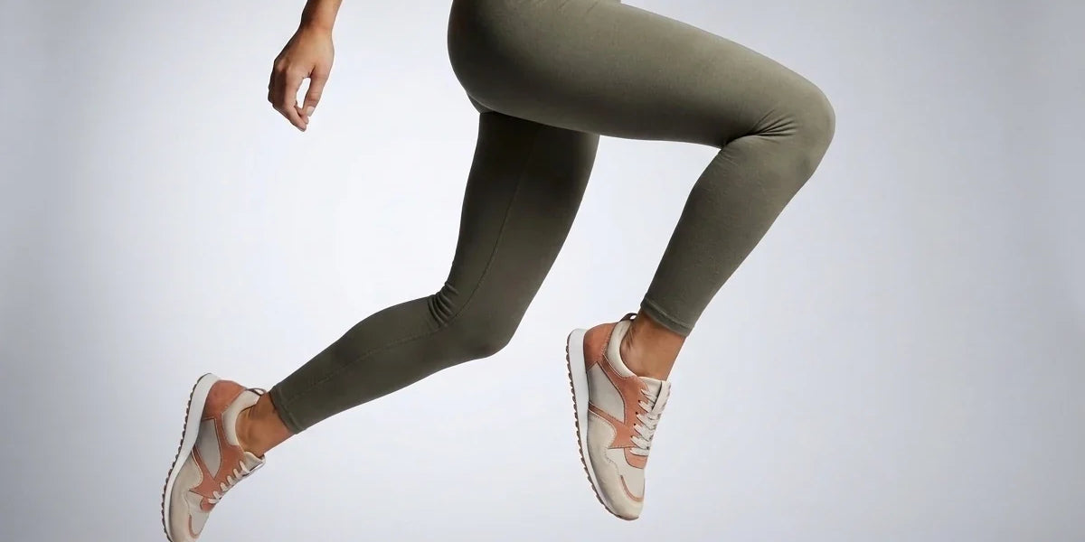 Side view of a woman mid-air wearing olive green high-waisted leggings and neutral athletic sneakers.