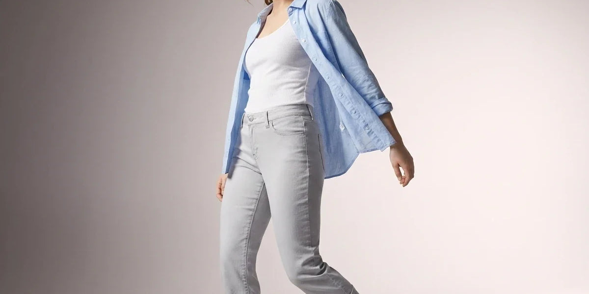 A woman modeling a light blue button-up shirt over a white tank top and light gray cropped jeans, standing against a neutral studio background.