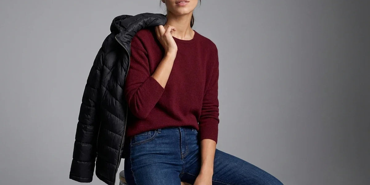 A woman seated on a chair against a gray studio background, wearing a burgundy knit sweater and blue jeans with a black puffer jacket draped over her shoulder.