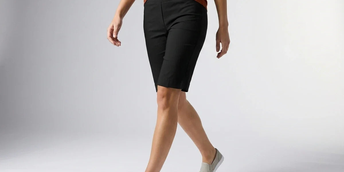 A model wearing black knee-length athletic shorts, a rust-orange top, and light gray slip-on sneakers, captured from the waist down against a light gray studio background.