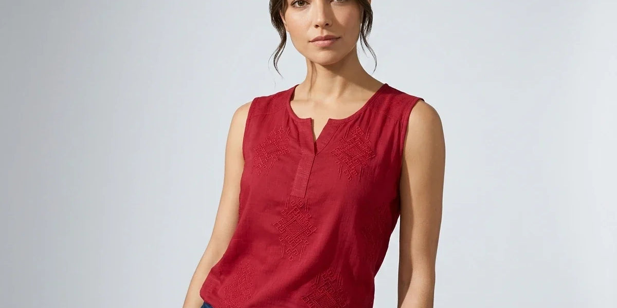 A woman modeling a sleeveless red top with a split neckline and tonal embroidered details, paired with blue jeans, against a light gray studio background.