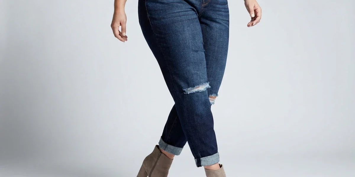 A model wearing dark blue distressed skinny jeans with cuffed hems, paired with a white shirt and tan ankle boots, standing against a light gray studio background.