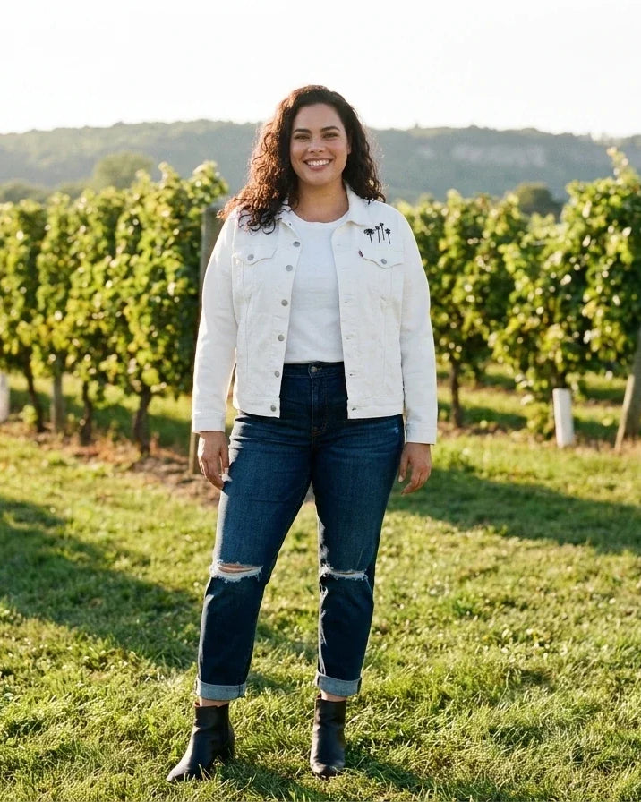 A woman stands in a sunny vineyard wearing a white denim jacket, distressed dark blue jeans, and black ankle boots.