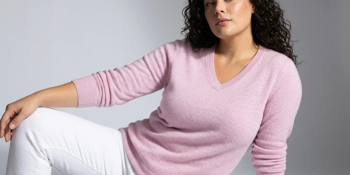 A woman with red hair wearing a light pink long-sleeve V-neck sweater and blue jeans, posing against a neutral light gray background.
