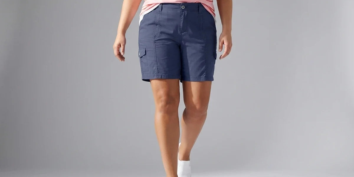 A model wearing navy blue cargo shorts, a pink and white striped t-shirt, and white socks walking against a light gray studio background.