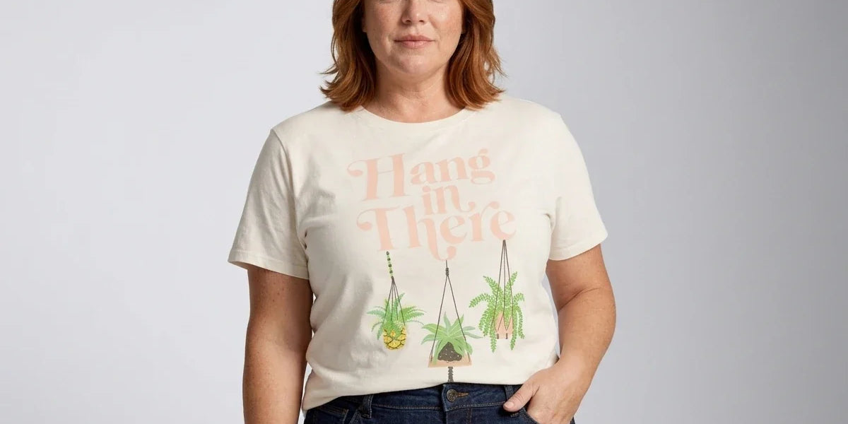 A woman with red hair wearing a cream-colored graphic t-shirt with the words 'Hang in There' and illustrations of hanging plants, paired with dark blue jeans, against a light gray studio background.