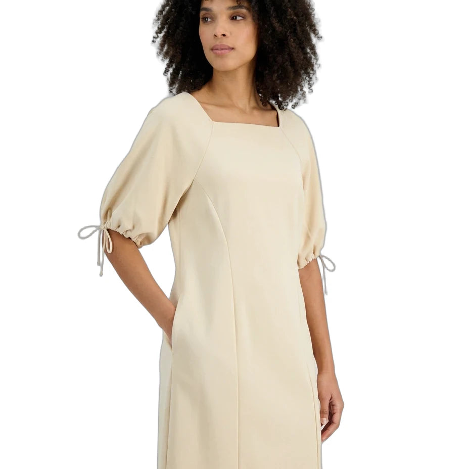 A woman with curly dark hair wearing a cream-colored dress with puff sleeves and tie details on the cuffs, set against a plain white background.