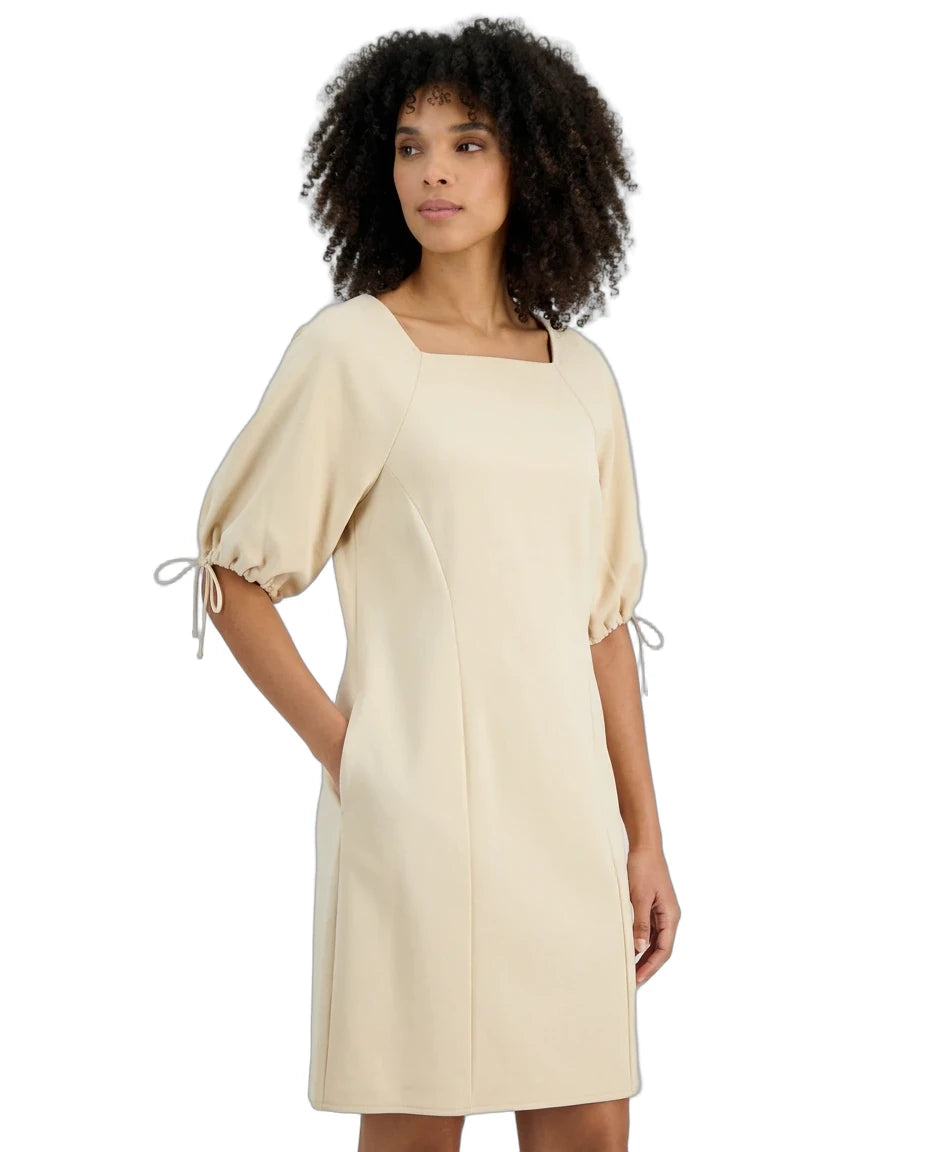 A woman with curly dark hair wearing a cream-colored dress with puff sleeves and tie details on the cuffs, set against a plain white background.