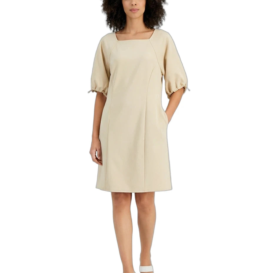 A woman modeling a light beige midi dress with puff sleeves, a square neckline, and side pockets, paired with white strappy sandals against a plain white background.