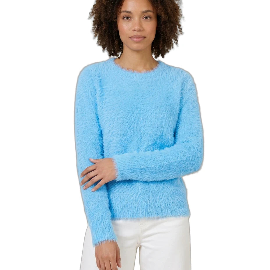 A woman with curly brown hair wearing a fuzzy light blue long-sleeve sweater and white pants, standing against a plain white background.