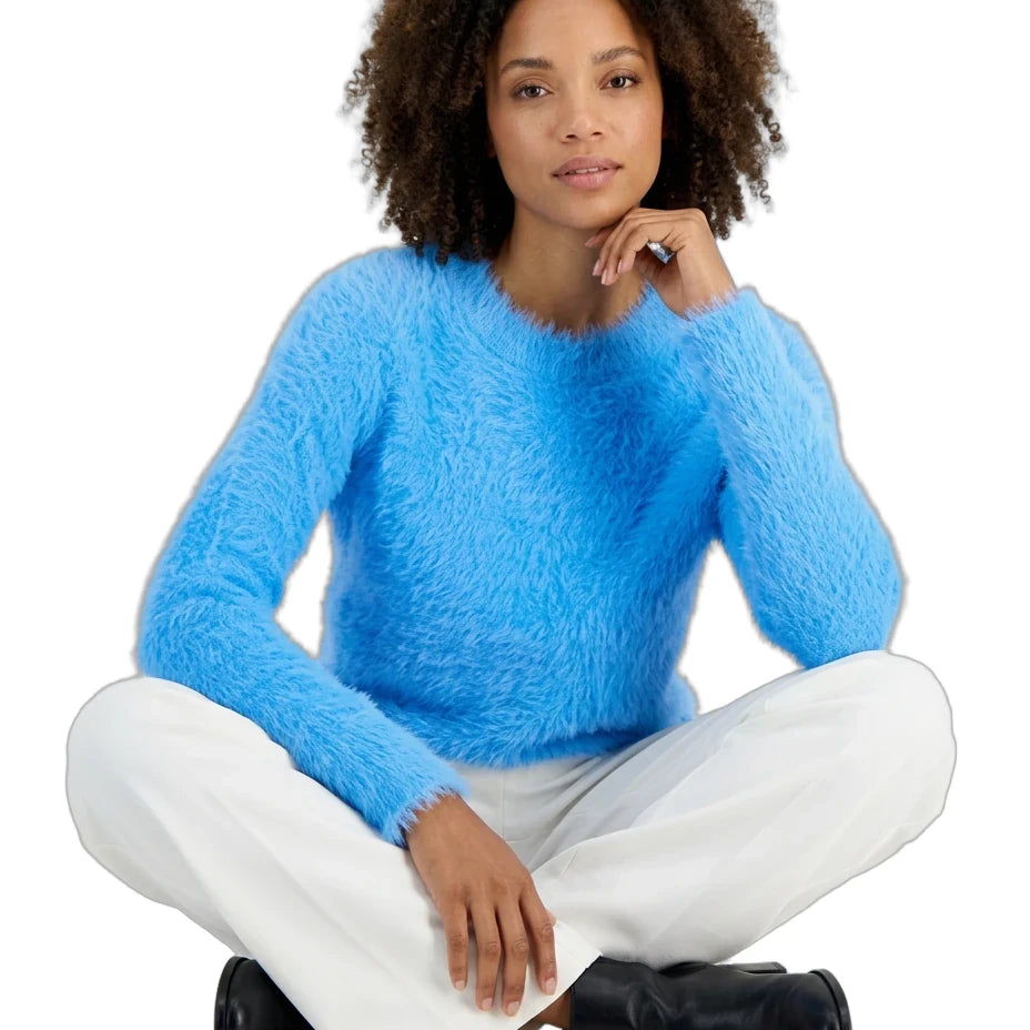 A woman with curly dark hair sits cross-legged against a white background, wearing a fuzzy bright blue long-sleeve sweater, white pants, and black boots.
