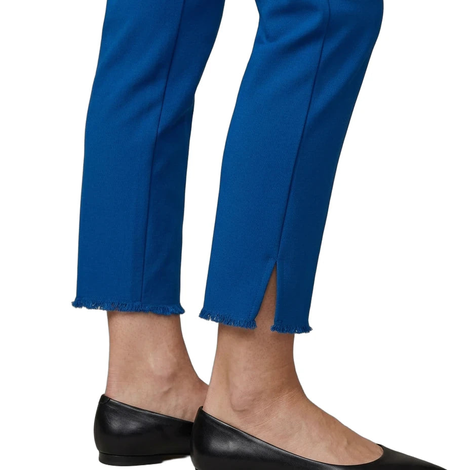 A close-up of a person's legs wearing bright blue cropped trousers with a raw frayed hem and side slits, paired with black pointed-toe flats, against a plain white background.