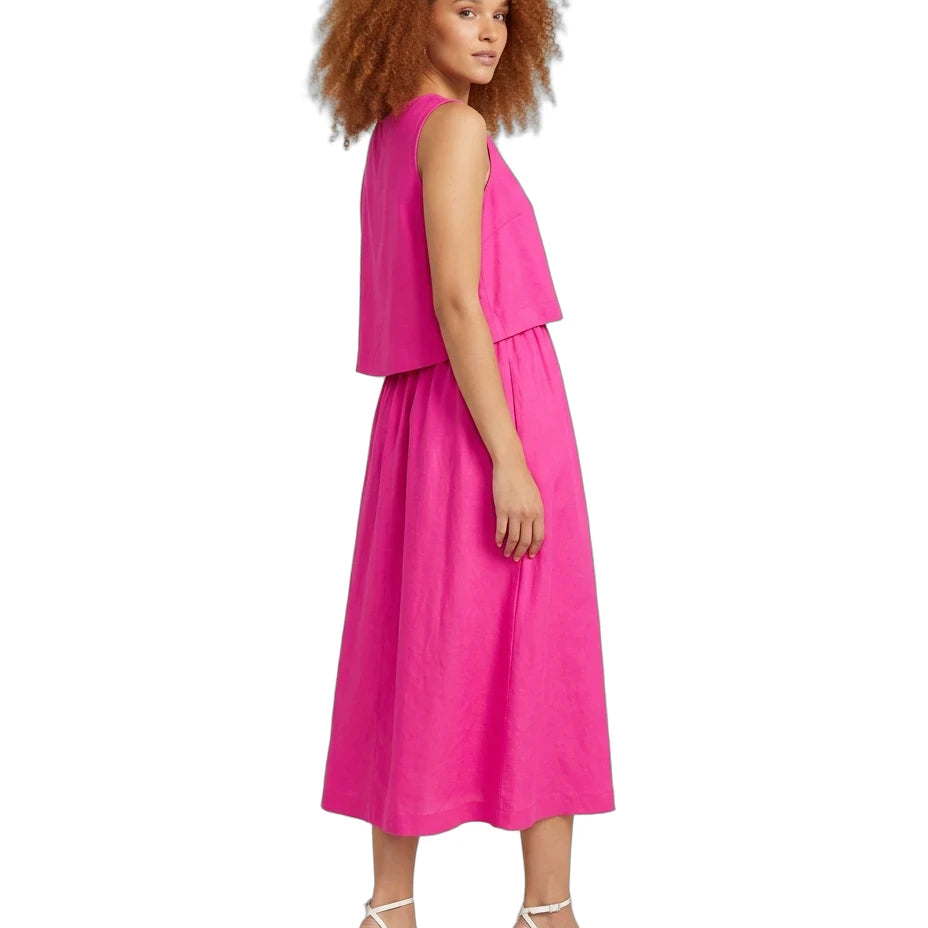 A woman with curly red hair is shown from the side wearing a bright pink sleeveless midi dress with a layered top and white strappy high-heeled sandals, set against a plain white background.