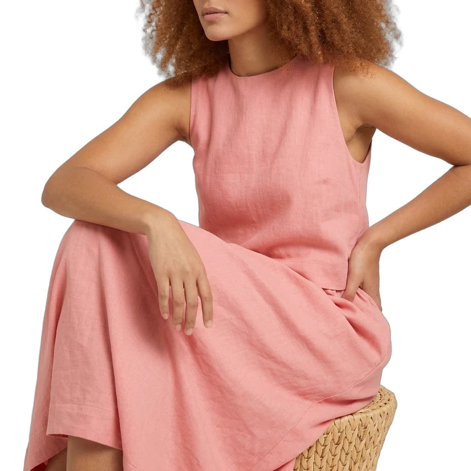 A woman with curly auburn hair wearing a sleeveless dusty pink linen dress, sitting on a woven straw chair against a white background.