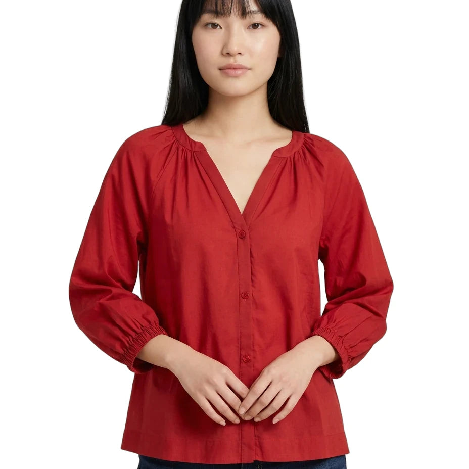 A woman wearing a red long-sleeve blouse with a split V-neckline, gathered shoulders, and button-front closure, paired with dark blue jeans against a plain white background.