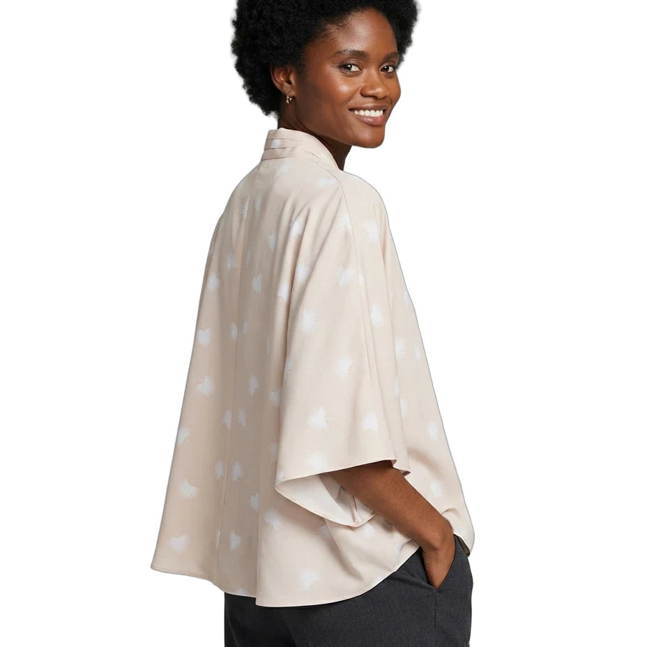 A woman with short black hair is shown from the back-three-quarter view wearing a light beige cape-style top with a subtle white star pattern, paired with dark gray pants, against a plain white background.