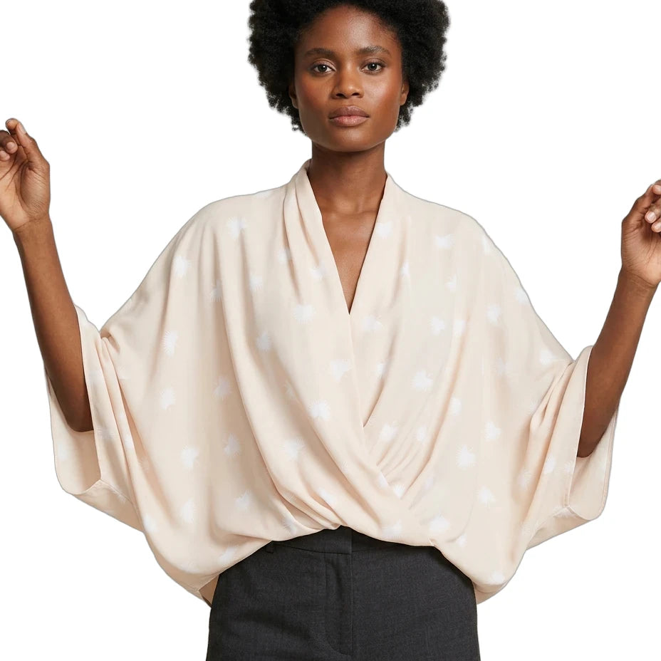 A woman modeling a light beige wrap-style blouse with a white dotted pattern and wide draped sleeves, paired with dark charcoal pants, against a plain white background.