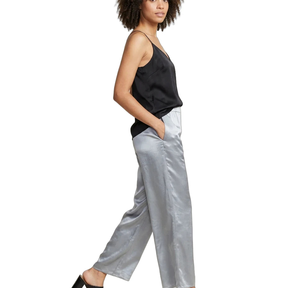 A woman modeling a black satin camisole and shiny silver satin wide-leg trousers, paired with black mule heels, against a plain white background.